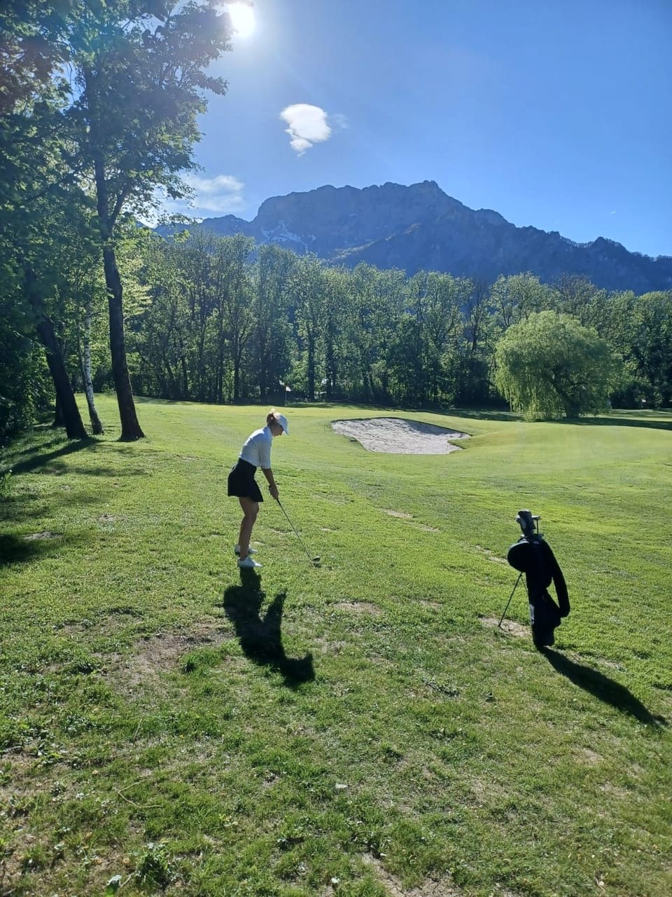 Anna in weißem Oberteil und Cap beim Abschlag auf einem sonnigen Golfplatz, dahinter ein Berg
