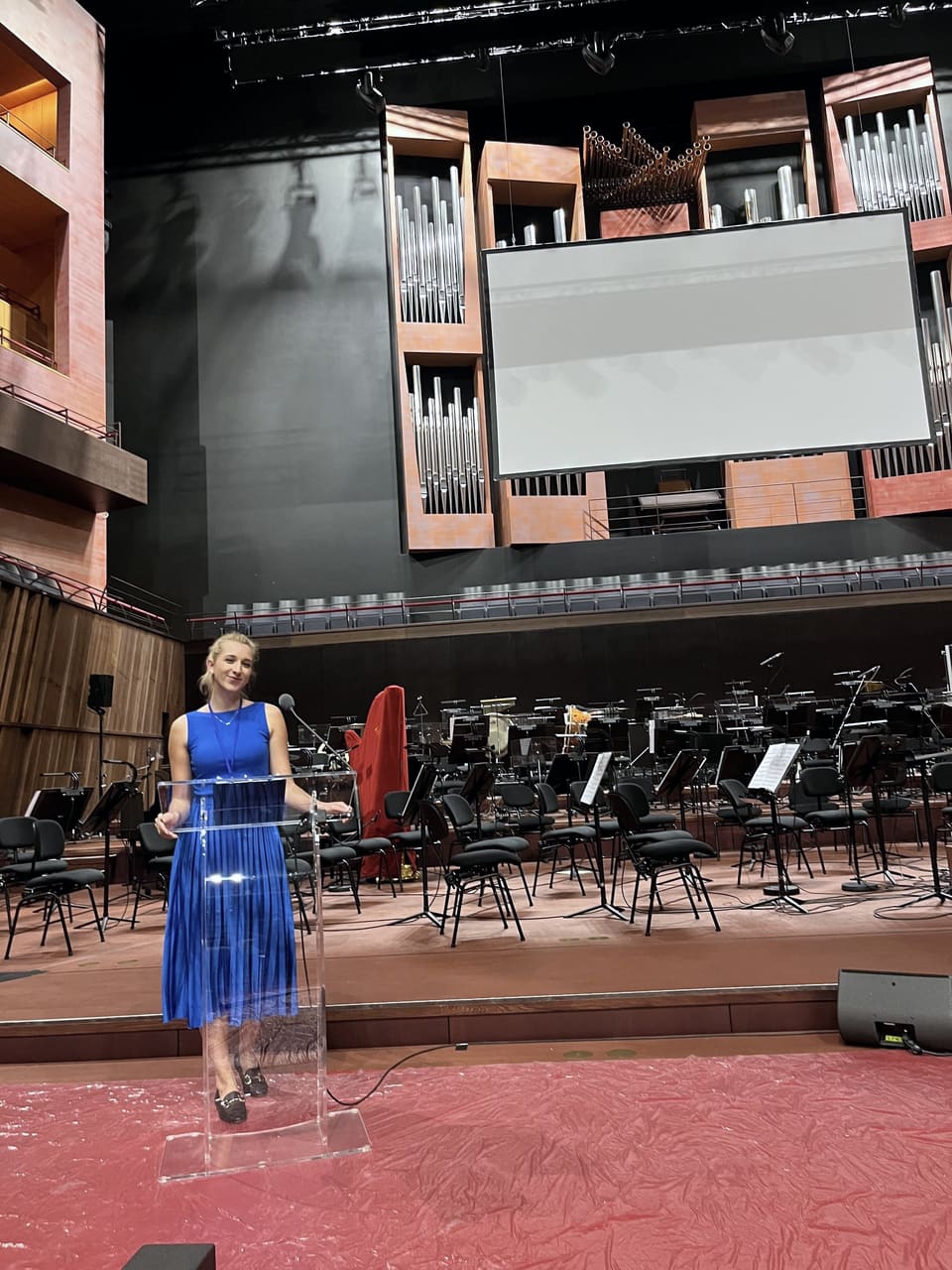 Anna in einem königsblauen Kleid mit Umhängeband, lächelnd hinter einem durchsichtigen Rednerpult auf einer leeren Konzertbühne mit einer Orgel dahinter