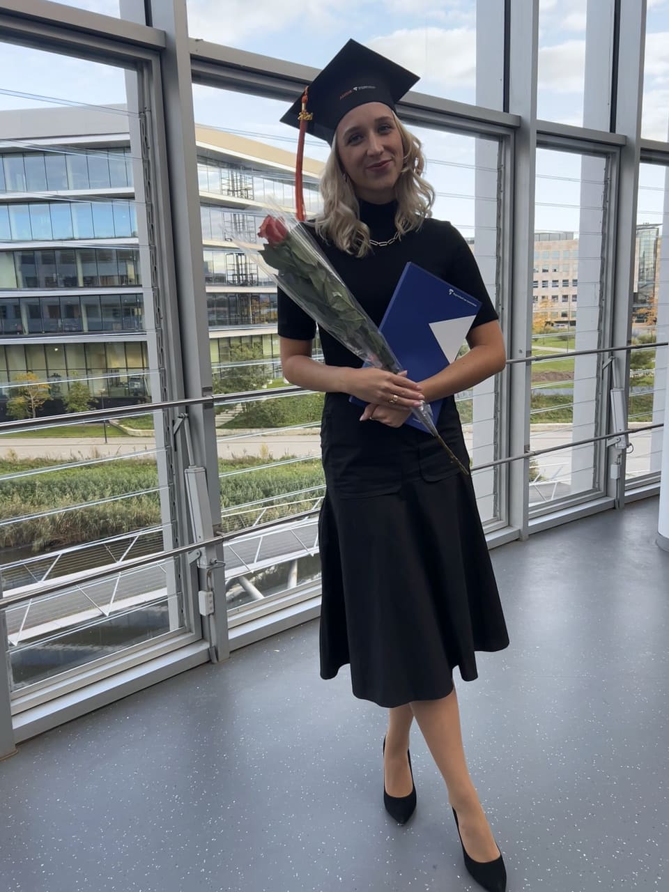 Anna mit Abschlusshut und schwarzem Kleid, mit einer roten Rose und einer blauen Mappe vor einer Glaswand