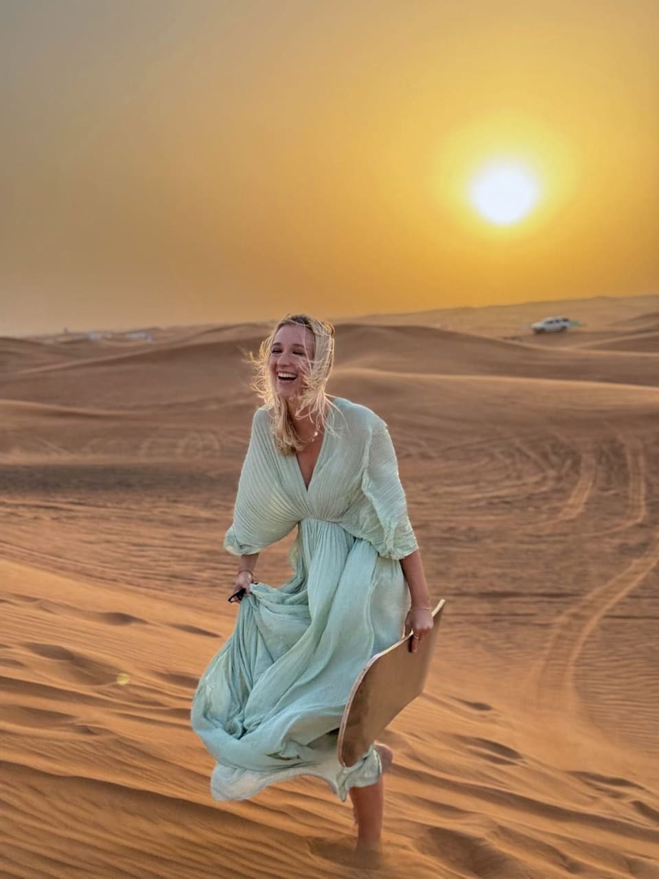 Anna lacht in einem fließenden hellgrünen Kleid auf einer Wüstendüne bei Sonnenuntergang und hält ein Holzbrett
