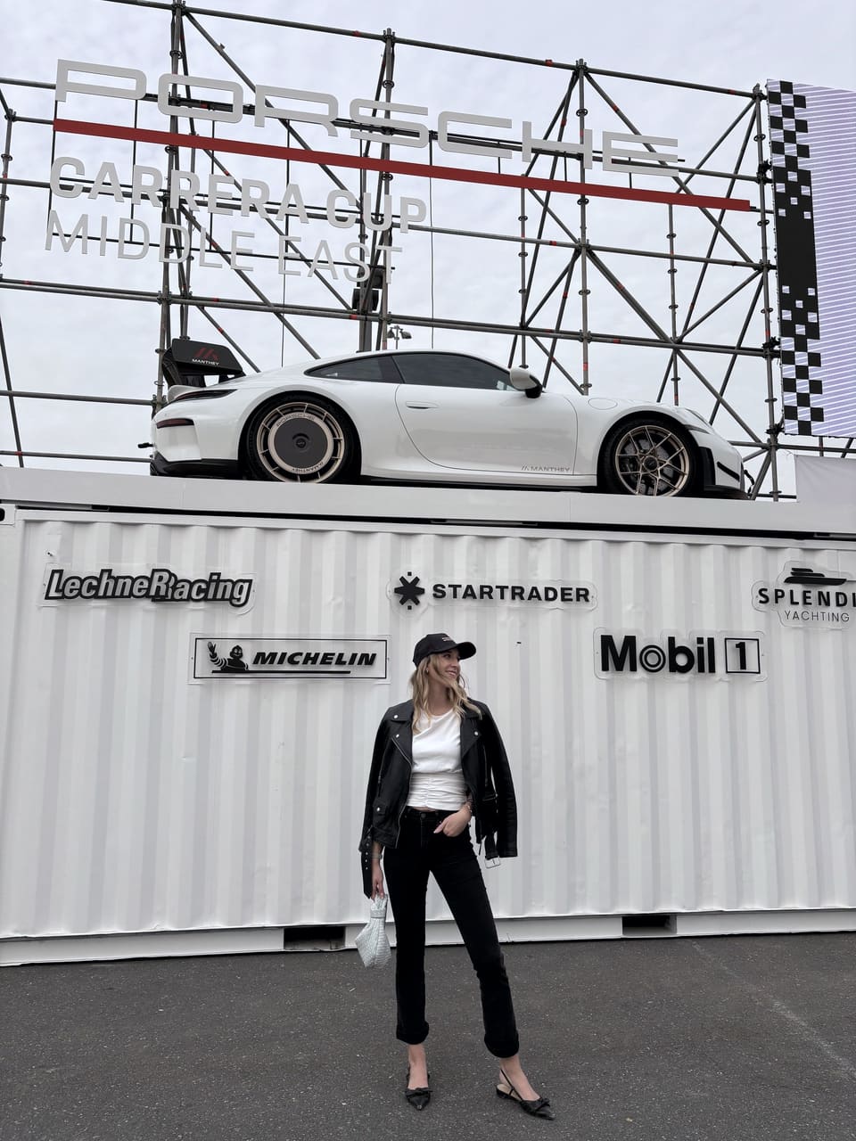 Anna in Cap und schwarzer Lederjacke vor einem weißen Sportwagen auf einem Schiffscontainer