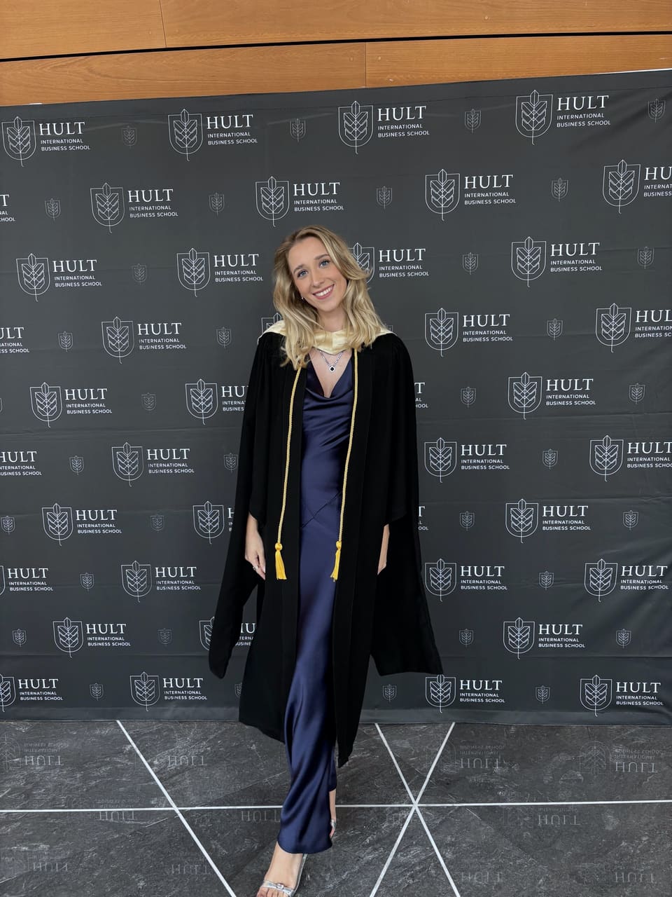 Anna lächelt in einem schwarzen Abschlussgewand mit goldenen Kordeln vor einer Schulwand