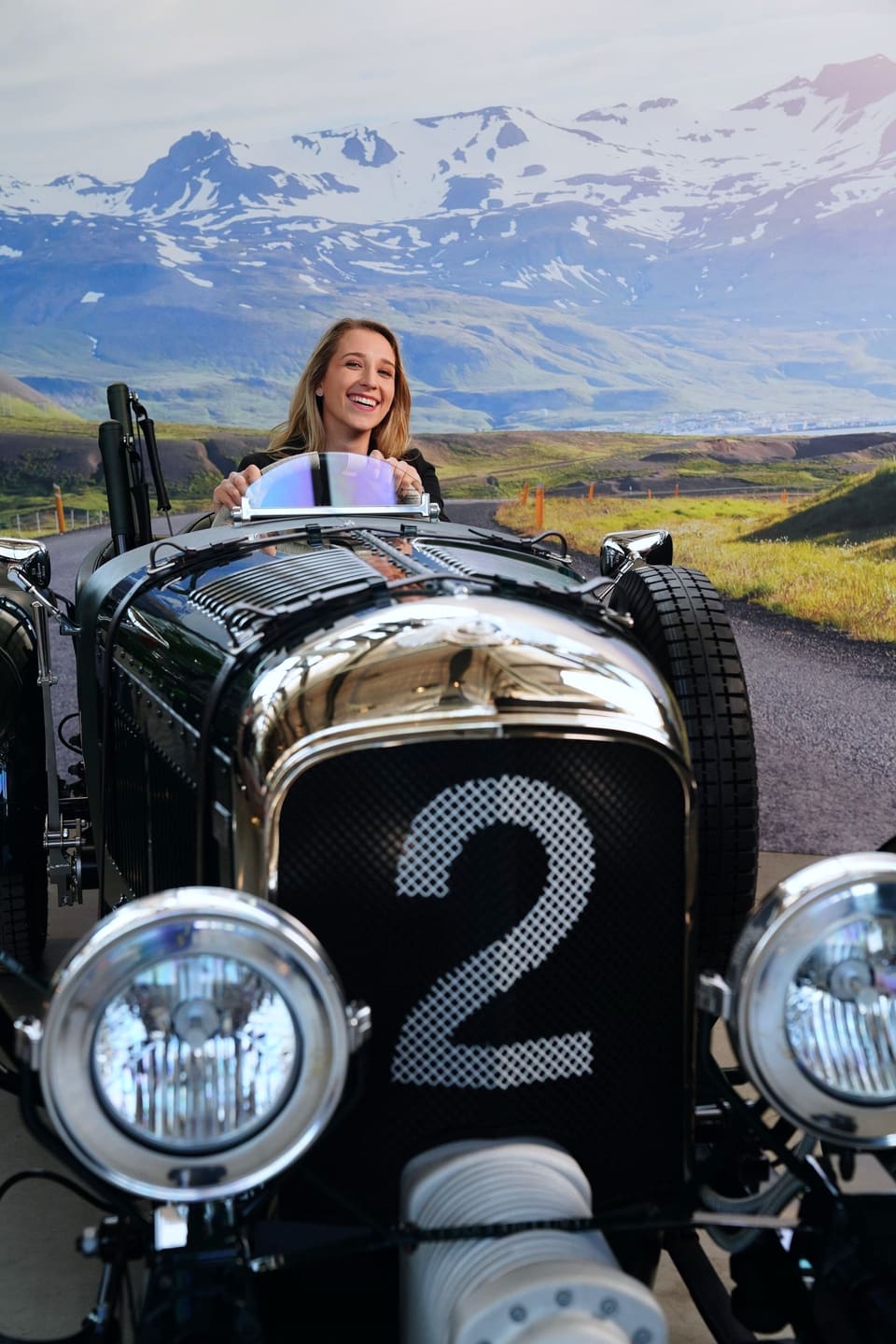 Anna lacht hinter der Windschutzscheibe eines Oldtimer-Rennwagens, dahinter ein Foto einer verschneiten Bergstraße