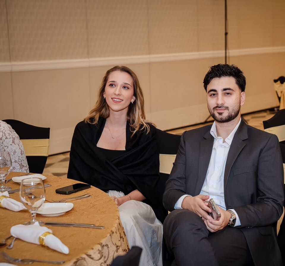 Anna lächelt an einem Bankettisch, neben ihr sitzt ein Mann im dunklen Anzug bei einem festlichen Abendessen